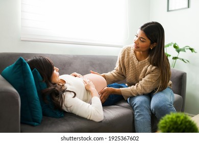 Hispanic midwife checking and touching the naked belly of a beautiful pregnant woman lying on the sofa in the living room