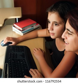 Hispanic Mother And Daughter Browsing The Web At Home With The Light From The Computer Monitor Illuminating Their Faces (with Books On The Computer Desk)