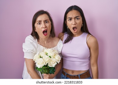 Hispanic Mother And Daughter Holding Bouquet Of White Flowers In Shock Face, Looking Skeptical And Sarcastic, Surprised With Open Mouth 