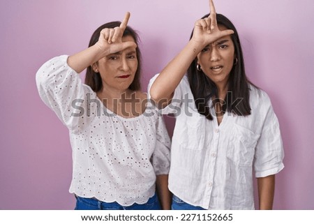 Hispanic mother and daughter together making fun of people with fingers on forehead doing loser gesture mocking and insulting. 