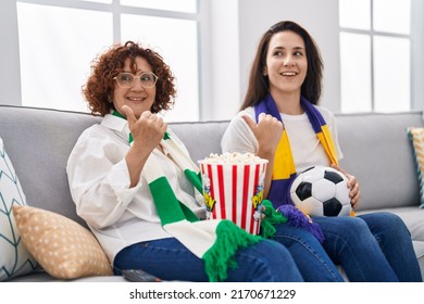 Hispanic Mother And Daughter Watching Football Supporting Team Pointing Thumb Up To The Side Smiling Happy With Open Mouth 