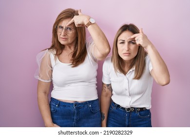 Hispanic Mother And Daughter Wearing Casual White T Shirt Over Pink Background Pointing Unhappy To Pimple On Forehead, Ugly Infection Of Blackhead. Acne And Skin Problem 