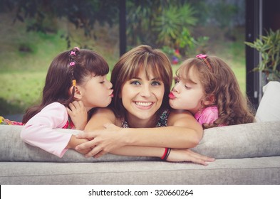 Hispanic Mother In Sofa With Two Daughters Kissing Her Cheeks From Each Side, Blurry Garden Background.