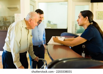 Hispanic Nurse Giving Admission Information About The Retirement Home To A Mature Old Man Using A Walker At The Nursery Home