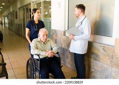 Hispanic Nurse Talking With The Doctor At The Retirement Home While Bringing A Sick Old Man In A Wheelchair At The Nursing Home