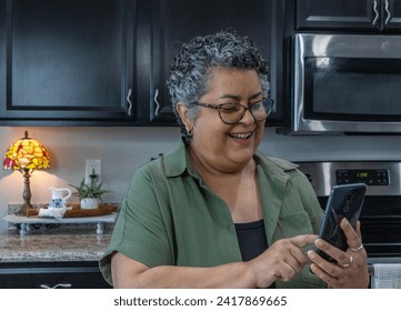 Hispanic older woman with curly gray hair is holding phone and smiling in kitchen, with copy space - Powered by Shutterstock