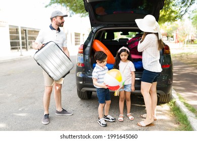 Hispanic Parents Packing Their Suitcases In The Trunk Of Their Car With Their Children. Young Mom, Dad And Little Kids Ready To Start A Family Vacation