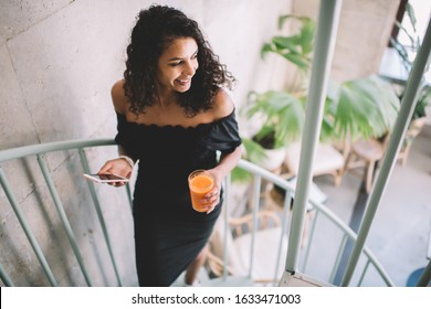 Hispanic Pleased Businesswoman In Elegant Black Dress With Glass Of Fresh Juice Smiling Away While Walking Up Stairs And Using Smartphone Against Blurred Lobby Interior Of Contemporary Building