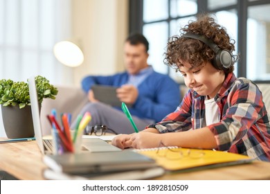 Hispanic School Boy Wearing Headphones, Preparing Homework While Sitting At The Desk At Home. Father And Son Spending Time Together