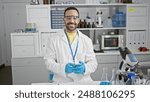 Hispanic scientist man with beard smiling in laboratory wearing lab coat and safety glasses, holding smartphone.