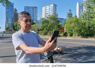 Hispanic Senior Man Looking At His Smart Phone Before Riding His Electric Kick Scooter In The City A Sunny Morning. Concepts Of Technology, Active Retired Life And Electrical Mobility.