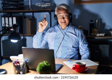 Hispanic Senior Man Wearing Call Center Agent Headset At Night Pointing Finger Up With Successful Idea. Exited And Happy. Number One. 