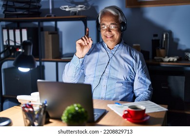 Hispanic Senior Man Wearing Call Center Agent Headset At Night Showing And Pointing Up With Finger Number One While Smiling Confident And Happy. 