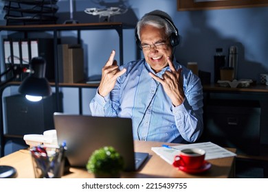 Hispanic Senior Man Wearing Call Center Agent Headset At Night Shouting With Crazy Expression Doing Rock Symbol With Hands Up. Music Star. Heavy Concept. 