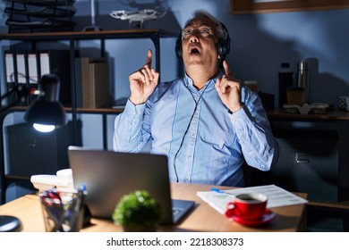 Hispanic Senior Man Wearing Call Center Agent Headset At Night Amazed And Surprised Looking Up And Pointing With Fingers And Raised Arms. 