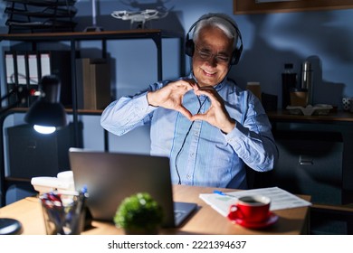 Hispanic Senior Man Wearing Call Center Agent Headset At Night Smiling In Love Doing Heart Symbol Shape With Hands. Romantic Concept. 