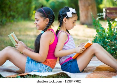 Hispanic Sisters Reading Book In Summer Park 