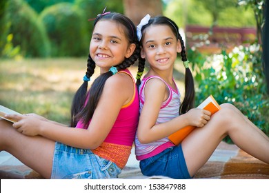 Hispanic Sisters Reading Book In Summer Park 