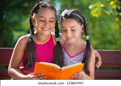 Hispanic Sisters Reading Book In Summer Park 