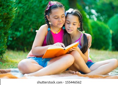 Hispanic Sisters Reading Book In Summer Park