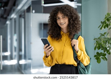 Hispanic student in modern hallway wearing yellow shirt and backpack, holding smartphone with confident smile. Image conveys education, technology, and contemporary lifestyle themes. - Powered by Shutterstock