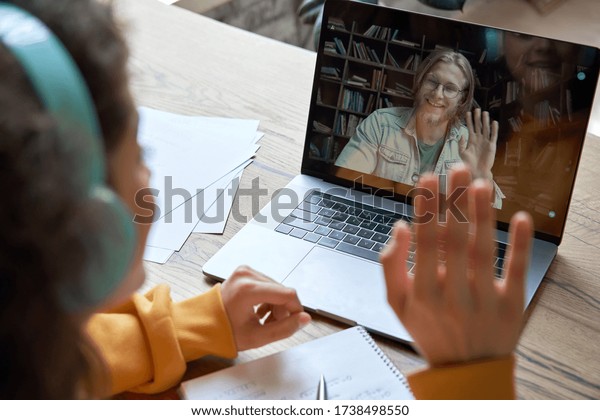 Hispanic teen girl school college student distance\
learning waving hand studying with online teacher on laptop screen.\
Elearning zoom video call, videoconference class with tutor. Over\
shoulder view.