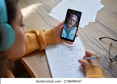 Hispanic teen girl school pupil college student wear headphones hold phone learning online with math teacher tutor using mobile video conference call app write in workbook. Over shoulder closeup view.