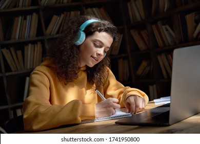 Hispanic Teen Girl School Pupil Or College Student Wearing Headphones Distance Elearning At Home Classroom Desk Writing Notes Watching Video Conference Call Remote Class Webinar Online On Computer.