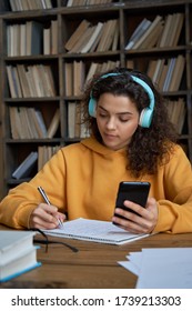 Hispanic teen girl school student wear headphones hold smartphone using distance learning mobile app online watching video course or zoom calling making notes in workbook sit at home or in library.