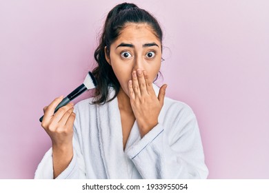 Hispanic Teenager Girl With Dental Braces Wearing Robe Holding Makeup Brush Covering Mouth With Hand, Shocked And Afraid For Mistake. Surprised Expression 