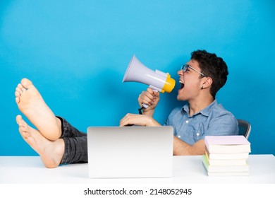 Hispanic teenager sitting feet on desk shouting on megaphone or speaker in front of laptop isolated on blue background.