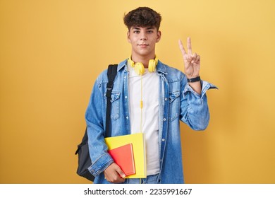 Hispanic teenager wearing student backpack and holding books smiling looking to the camera showing fingers doing victory sign. number two. 
