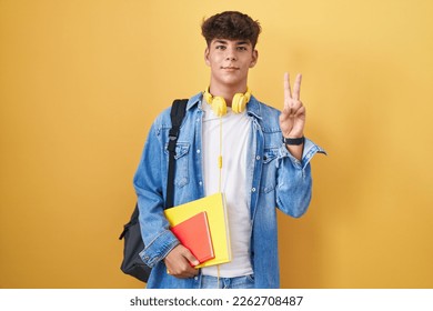 Hispanic teenager wearing student backpack and holding books smiling with happy face winking at the camera doing victory sign with fingers. number two. 