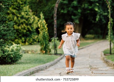 Hispanic Toddler Girl Two Years Old In White Polka-dot Shirt And Blue Denim Shorts Running On Track Of Garden Or Park.