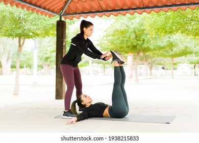 Hispanic Woman In Activewear Helping Her Workout Partner To Do Leg Raise Abs Exercise To Strengthen The Abdomen. Two Women Training In The Park