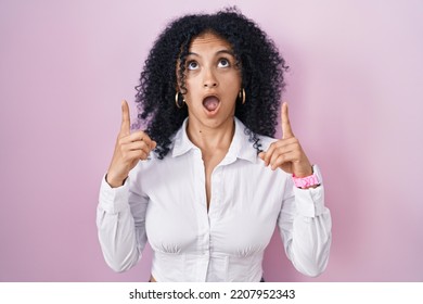 Hispanic Woman With Curly Hair Standing Over Pink Background Amazed And Surprised Looking Up And Pointing With Fingers And Raised Arms. 