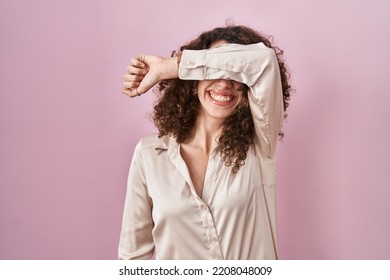 Hispanic Woman With Curly Hair Standing Over Pink Background Covering Eyes With Arm Smiling Cheerful And Funny. Blind Concept. 