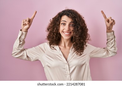 Hispanic Woman With Curly Hair Standing Over Pink Background Smiling Amazed And Surprised And Pointing Up With Fingers And Raised Arms. 