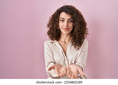 Hispanic woman with curly hair standing over pink background smiling with hands palms together receiving or giving gesture. hold and protection  - Shutterstock ID 2254802189