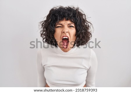 Hispanic woman with curly hair standing over isolated background angry and mad screaming frustrated and furious, shouting with anger. rage and aggressive concept. 