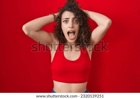Hispanic woman with curly hair standing over red background crazy and scared with hands on head, afraid and surprised of shock with open mouth 