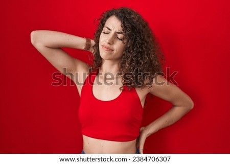 Hispanic woman with curly hair standing over red background suffering of neck ache injury, touching neck with hand, muscular pain 