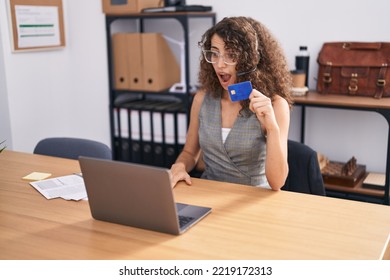 Hispanic Woman With Curly Hair Wearing Call Center Agent Headset Holding Credit Card Scared And Amazed With Open Mouth For Surprise, Disbelief Face 