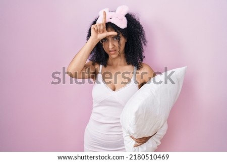 Hispanic woman with curly hair wearing sleep mask and pajama holding pillow making fun of people with fingers on forehead doing loser gesture mocking and insulting. 