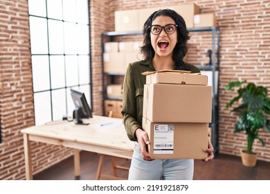 Hispanic Woman With Dark Hair Working At Small Business Ecommerce Holding Boxes Angry And Mad Screaming Frustrated And Furious, Shouting With Anger Looking Up. 