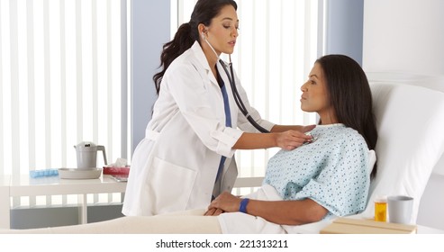 Hispanic Woman Doctor Listening To Patient's Heart In Hospital Bed