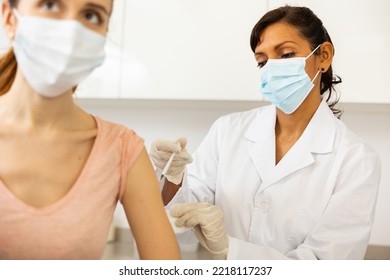 Hispanic Woman Doctor Vaccinating Young Caucasian Woman Patient. They're Wearing Face Masks.