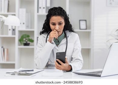 A Hispanic woman, dressed as a doctor, appears anxious while looking at a text message on her smartphone in a medical office setting. - Powered by Shutterstock