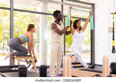 Hispanic Woman Exercising With Pilates Band, Young European Woman Working Out On Pilates Chair. African-american Man Instructor Assisting Them With Workout.