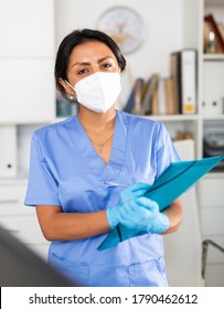 Hispanic Woman General Practitioner In Blue Uniform And Face Mask Standing In Clinic, Filling Out Clipboard With Medical Records